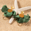 Les boucles d'oreilles Dounia : des boucles d'oreilles en tissu en forme de fleur vertes. Elles sont également constituées de pendentif rond en métal doré d'un flash d'or 24 carats et d'un anneau ovale en métal doré d'un flash d'or 24 carats.