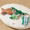 Porte-clefs Adibou : composé d'un poisson en bois, d'un cordon en cuir vert, d'une perle en verre recyclé verte et d'un médaillon en forme de barre de navire en plaqué argent 999.