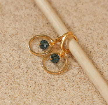 Boucles d’oreilles Siloe dorées avec perles bleu gris posées sur sable – fabrication artisanale française