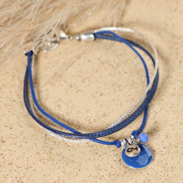 Bracelet Holiday argenté bleu marine posé à plat, montrant ses fils tressés bleu marine, argent et bleu, avec pendentif en forme de poisson en métal plaqué argent 999, perle en verre bleu marine et médaille Retour de Plage