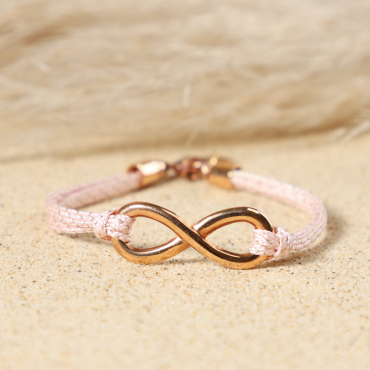 Gros plan sur le pendentif infini en métal doré rose du bracelet Infini, entouré de cordon en coton rose et médaille Retour de Plage, fabrication artisanale sur l'île d'Oléron