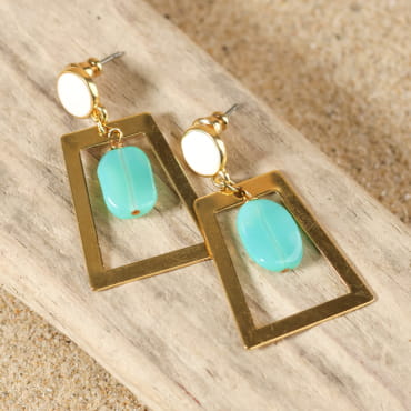 boucles d’oreilles Niamey dorées avec pendentif rectangulaire et perle verte menthe présentées sur fond sable