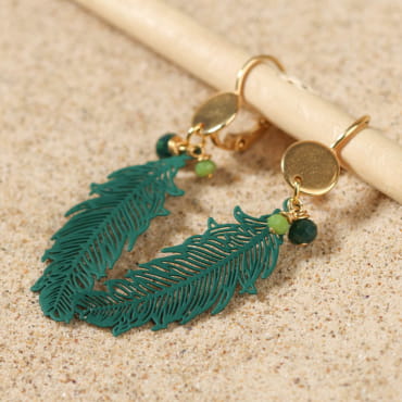 Boucles Ara dorées à plumes vertes, inspiration exotique, posées sur fond sable – fabrication française