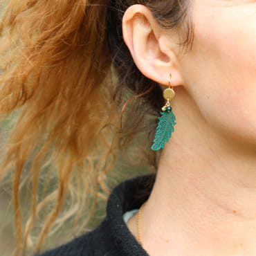 Boucles d’oreilles pendantes dorées et vertes, en forme de plumes stylisées, portées à l’oreille.