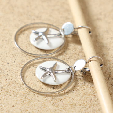 Gros plan sur les boucles d’oreilles Babette argentées, mettant en valeur le pendentif émaillé blanc rond, l’étoile de mer argentée et l’anneau circulaire, fabrication artisanale sur l'île d'Oléron – Retour de Plage