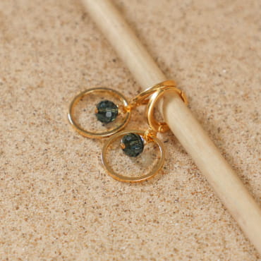 Boucles d’oreilles Siloe dorées avec perles bleu gris posées sur sable – fabrication artisanale française