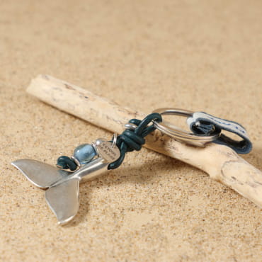 Gros plan sur le pendentif en forme de queue de baleine en métal plaqué argent 999 du porte-clés Arcachon bleu ciel, accompagné de perle en verre bleu transparent et perle argentée