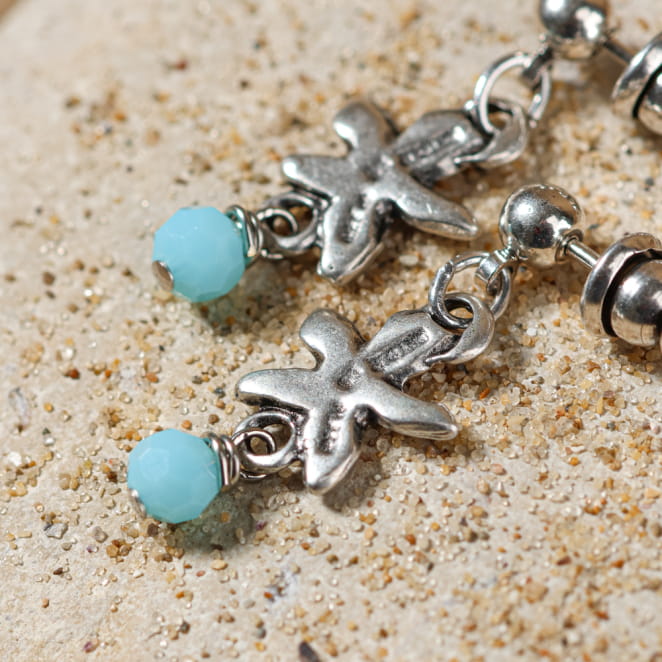 Boucles d’oreilles Ibiza enfant bleu ciel et argent, posées sur le sable – fabrication artisanale française