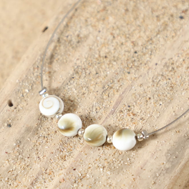 Détail des perles en forme d'œil de Sainte-Lucie et des petites perles en métal plaqué argent 999 du bracelet Ajaccio, sur câble en acier argenté, fermoir mousqueton et médaille Retour de Plage.