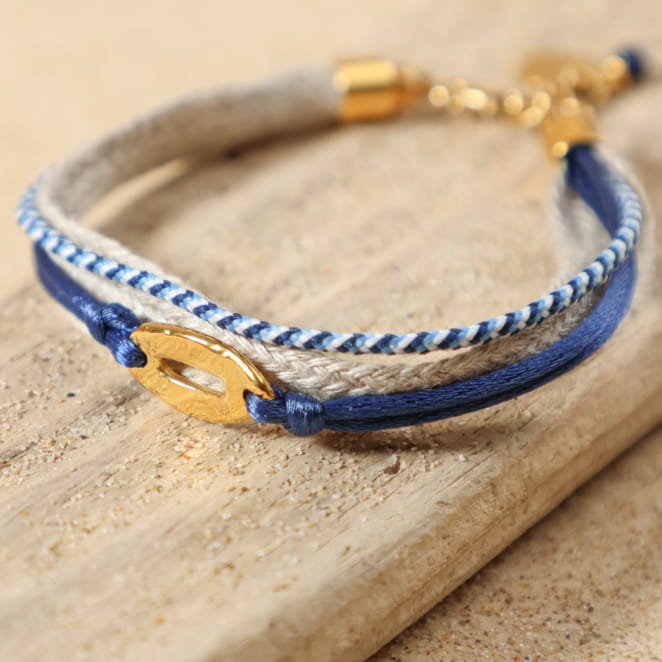 Bracelet Chenal doré bleu marine posé à plat, montrant ses cordons en lin naturel, coton bleu et blanc, et bleu marine satiné, avec pendentif ovale en métal doré à l'or fin 24 carats, fermoir mousqueton et médaille Retour de Plage.