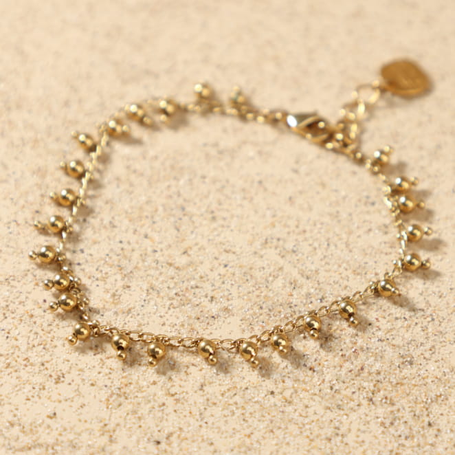 Bracelet Grelot doré posé à plat, montrant sa chaîne délicate ornée de petites perles rondes en métal doré 24 carats, avec médaille Retour de Plage, fait main en France.