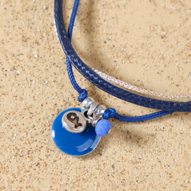 Gros plan sur le pendentif en forme de poisson en métal plaqué argent 999 du bracelet Holiday, accompagné d'une perle en verre bleu marine et de fils tressés bleu marine, argent et bleu, avec médaille Retour de Plage.
