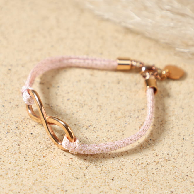 Bracelet Infini doré rose posé à plat, montrant son cordon en coton rose, pendentif infini en métal doré rose et médaille Retour de Plage, fait main en France