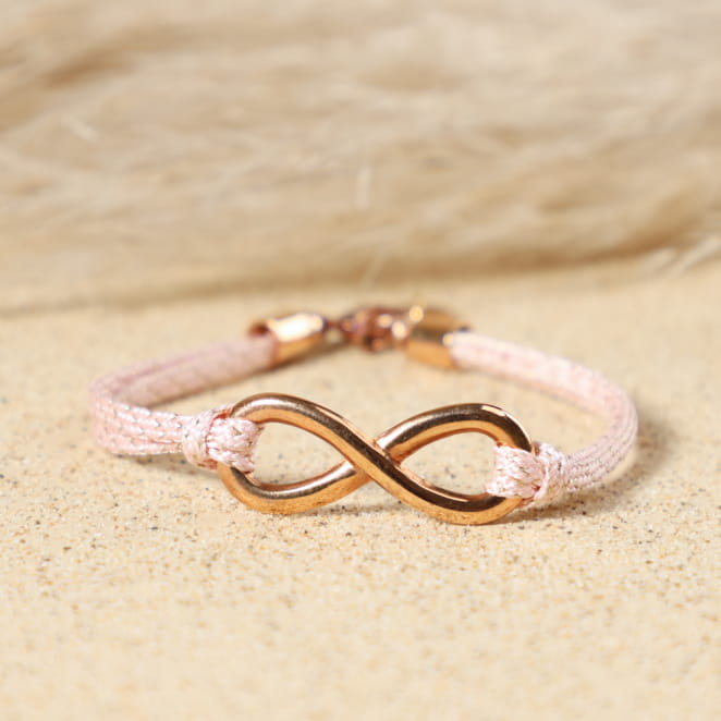 Gros plan sur le pendentif infini en métal doré rose du bracelet Infini, entouré de cordon en coton rose et médaille Retour de Plage, fabrication artisanale sur l'île d'Oléron