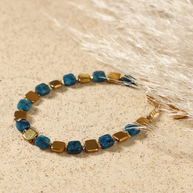Bracelet Solal doré bleu pétrole posé à plat, montrant son design élégant avec perles carrées et médaille Retour de Plage, sur câble fin doré à l'or fin.
