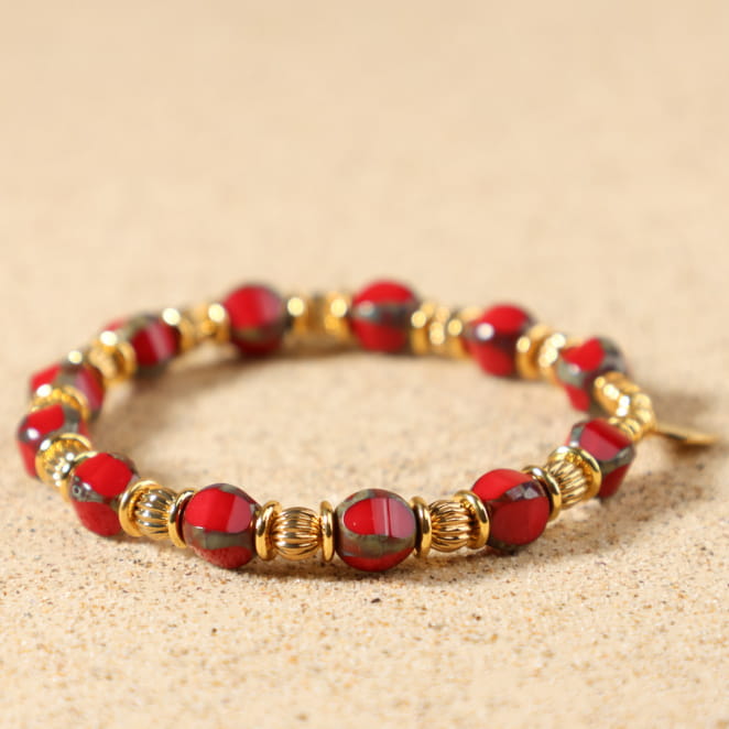 Gros plan sur les perles en verre rouge et les perles dorées finement texturées du bracelet Tequila, sur fil élastique transparent, médaille Retour de Plage