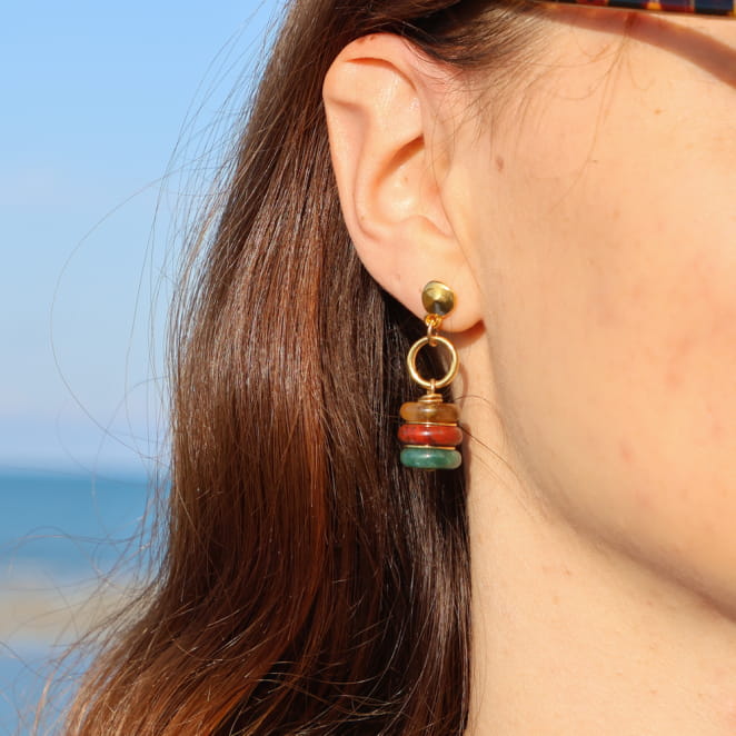 boucles d’oreilles Giens dorées avec pendentif blanc présentées sur fond sable