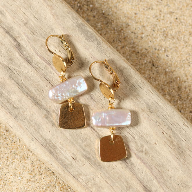 boucles d’oreilles Giens dorées avec pendentif blanc présentées sur fond sable