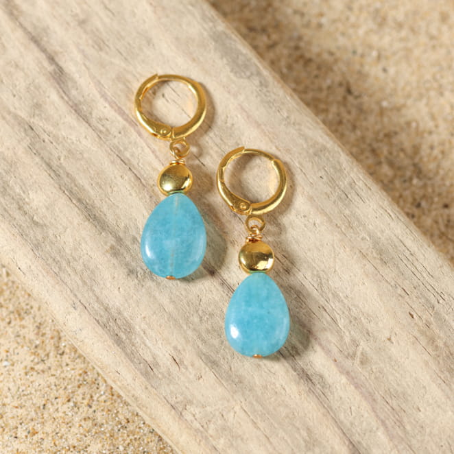 boucles d’oreilles Mombasa dorées avec pendentif bleu et perle turquoise présentées sur fond sable