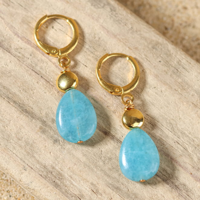 boucles d’oreilles Mombasa dorées avec pendentif bleu et perle turquoise présentées sur fond sable