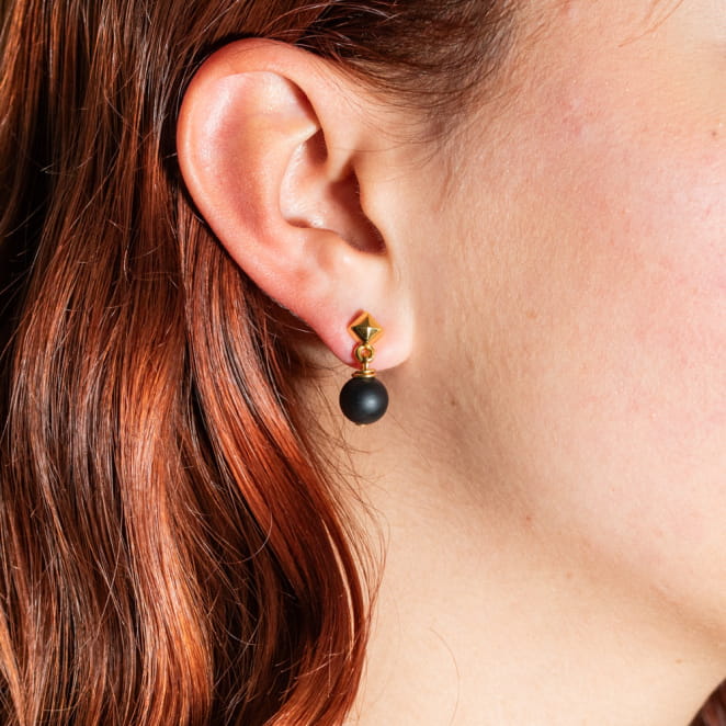 Atome est une paire de petites boucles d'oreilles composée d'un clou d'oreilles en métal doré d'un flash d'or 24 carats et d'un pendentif boule noir. Il s'agit d'une pièce unique fabriquée sur l'ïle d'Oléron.