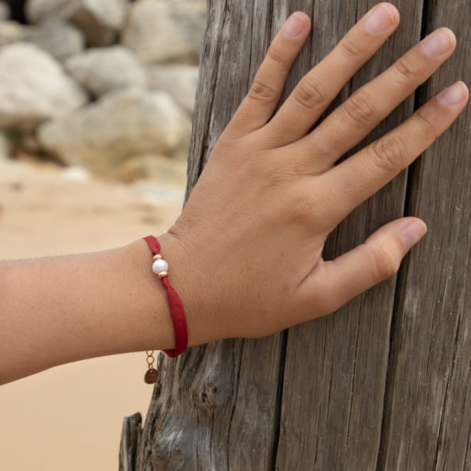 Le bracelet Céline : une pièce unique composé nd'un ruban en polyester rouge cerise, d'une perle de culture de rivière blanche, de perles en métal doré rose et d'une médaille Retour de plage doré rose. Ce bijou ajoute couleur et légèreté à votre tenue.