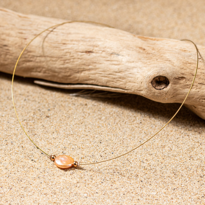 collier Flamand doré avec pendentif ovale rose saumon posé sur un bois flotté