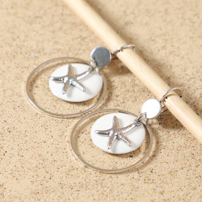 Boucles d’oreilles Babette argentées posées à plat, montrant leur design élégant avec pendentif émaillé blanc rond, étoile de mer argentée et anneau circulaire, fait main en France – Retour de Plage