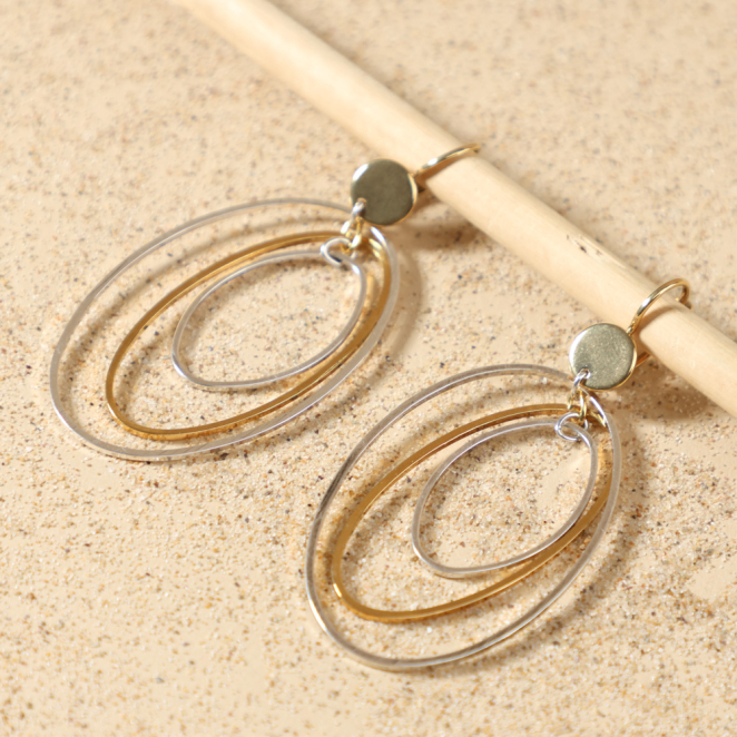 Gros plan sur les boucles d’oreilles Proton, montrant les trois anneaux ovales imbriqués en métal plaqué argent et doré à l'or fin 24 carats, fabrication artisanale sur l'île d'Oléron – Retour de Plage
