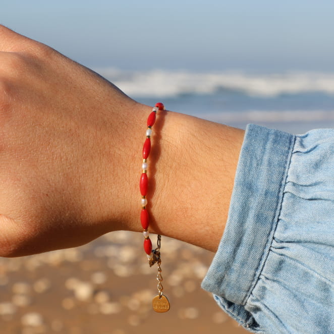 Bracelet doré à perles rouges intenses, porté sur fond de sable clair.