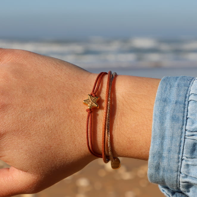 Bracelet doré à perles marron, finition brillante, porté sur poignet au bord de l’eau.