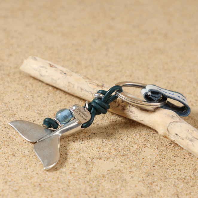 Gros plan sur le pendentif en forme de queue de baleine en métal plaqué argent 999 du porte-clés Arcachon bleu ciel, accompagné de perle en verre bleu transparent et perle argentée
