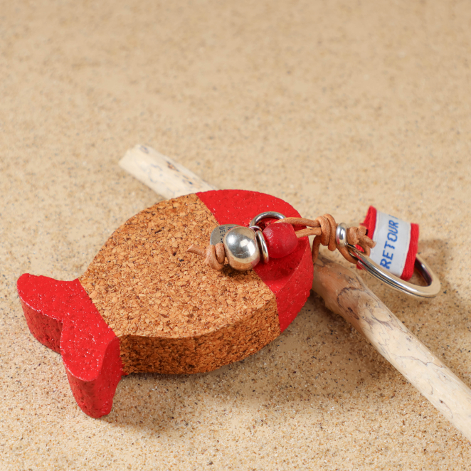 Gros plan sur le flotteur en forme de poisson en liège du porte-clés Fletan rouge, avec perle en céramique rouge, perle en métal plaqué argent 999, ruban en tissu Retour de Plage et médaille Retour de Plage argentée.